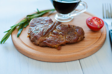 Cooked, grilled pork steak on a wooden round board, with soy sauce, rosemary and tomato, on light background with copy space