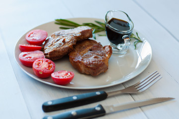 Tasty, grilled pork steaks on a gray plate with sauce, rosemary and tomatoes, on light wooden background, cutlery, soft focus