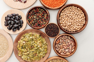 top view of wooden bowls with diverse beans, quinoa, red lentil, peppercorns, pumpkin seeds and chickpea on white marble surface