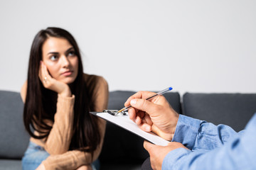 Young lady having depression sitting on the couch opposite to psychotherapist and crying. Male doctor holding clipboard intending to fill in all required information about medical patient
