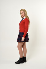 full length portrait of blonde girl wearing red cardigan with tie and plaid skirt, school uniform, side profile, standing  pose, on a white studio background.