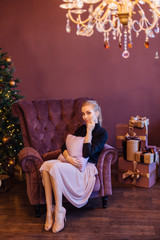 Beautiful young woman dressed in a sweater and skirt sitting on the arm chair next to the christmas tree with a pillow in hands.