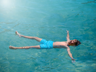 European boy is laying in blue clear sea water in form of sea star. He is relaxing during his...