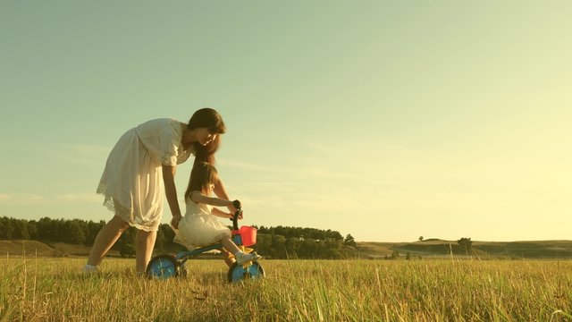 Happy Mother Teaches Little Daughter To Ride Bike. Mom Plays With Small Child On Lawn. Kid Learns To Ride Bicycle. Parents And Little Daughter Walks In Park. Concept Of Happy Family And Childhood.