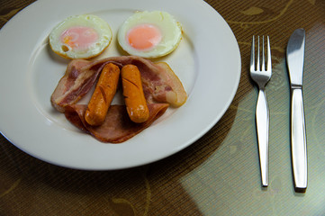 Yummy American breakfast with sunny fried eggs, rosted bacon, ham, two sausages in white dish with silver fork and knife on table.