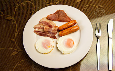 Yummy American breakfast with sunny fried eggs, rosted bacon, ham, two sausages in white dish with silver fork and knife on table.