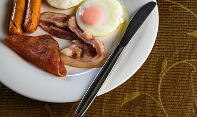 Closeup yummy American breakfast with sunny fried eggs, rosted bacon, ham, two sausages in white dish with silver knife on table.