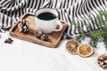 Winter composition. Breakfast in bed. Cup of coffee on wooden tray, wool blanket, fir tree branch, orange slices and pine cones. White linen table background. Festive cozy design. Selective focus. - Powered by Adobe
