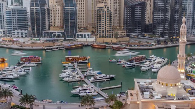 Yachts in Dubai Marina flanked by the Al Rahim Mosque and residential towers and skyscrapers aerial day to night transition timelapse. Modern high rise skyline with boats and wooden dhows