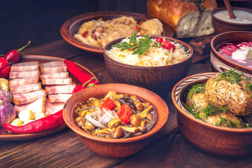 Traditional Ukrainian cuisine - canned mushrooms, baked potatoes, lard, hot peppers, sauerkraut, borsch, dumplings and donuts in clay pots on a dark wooden background.