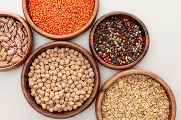 top view of wooden bowls with chickpea, oatmeal, red lentil, beans and peppercorns on white marble surface