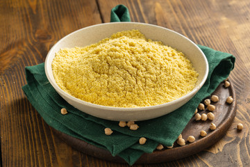 Chickpea flour in white ceramic bowl on wooden table with spilled chickpeas