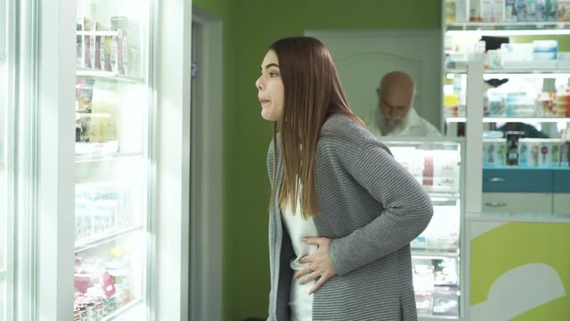 Young caucasian brunette girl with severe pain in the stomack staying at the pharmacy and choosing the pain medication. Senior pharmacist on the background waiting for his customer. Healthcare.