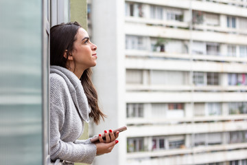 Young woman looking out the window at Sao Paulo city and holding cell phone. Thoughts about the...