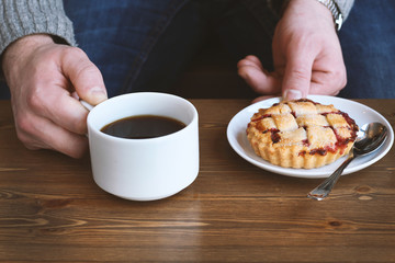 man in grey sweater drinks black lungo americano coffee in winter autumn in coffee shop eating classic american home-made cherry pie on white plate on wooden table. Fall mood instagram style