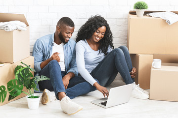 Cheerful couple sitting on floor at new apartment, using laptop