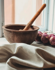 rustic flat lay, clay mortar with wooden pestle for grinding spices on linen background and string of onions on windowsill