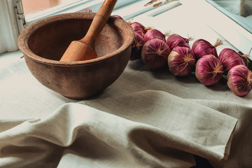rustic flat lay, clay mortar with wooden pestle for grinding spices on linen background and string of onions on windowsill