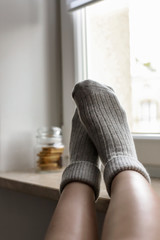 Women wearing grey knitted cozy socks on the windowsill, with cookies and a mug on the background of. A cozy home atmosphere.