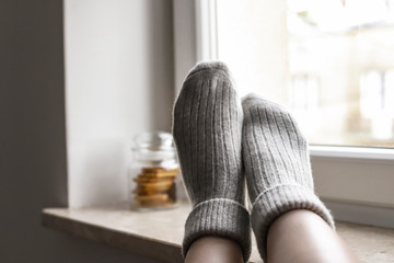 Women wearing grey knitted cozy socks on the windowsill, with cookies and a mug on the background of. A cozy home atmosphere.