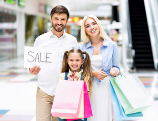 Family Holding Shopping Bags And Sale Sign Standing In Mall