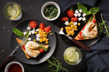 Two eggs plowed on toast with pieces of cheese, tomatoes, herbs and corn grains on a black background. Top view