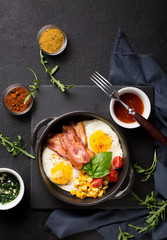 Fried egg with slices of bacon and spinach leaves in a black pan with handles on a black background. Classic English breakfast. Top view with copy space for your text