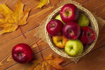 Red apples in a small basket and maple leaves on a wooden background, top view