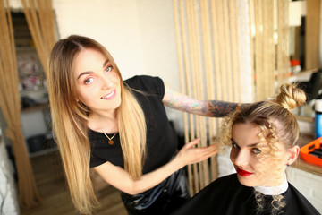 Hairdresser Styling Curls and Bun Girl Hairstyle. Professional Girl Hairstylist Making Curly Hairdo for Female Client. Stylist Showing Wavy Hair. Two Smiling Blonde Woman Looking at Camera Shot