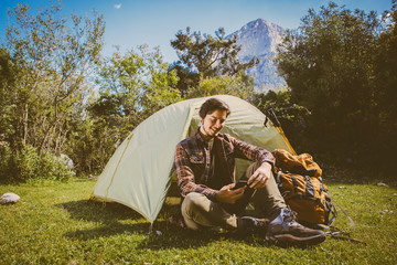 Young camper man is using smartphone with a smile on his face in front of the tent. Young camper man pitched his tent between trees on the green grass with a wonderful huge mountain view.