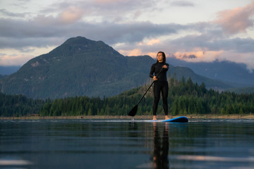 Fototapeta na wymiar Adventurous Girl on a Paddle Board is paddling in a calm lake with mountains in the background during a colorful summer sunset. Taken in Stave Lake near Vancouver, BC, Canada.