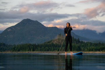 Adventurous Girl on a Paddle Board is paddling in a calm lake with mountains in the background during a colorful summer sunset. Taken in Stave Lake near Vancouver, BC, Canada.