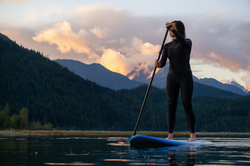 Adventurous Girl on a Paddle Board is paddling in a calm lake with mountains in the background during a colorful summer sunset. Taken in Stave Lake near Vancouver, BC, Canada.