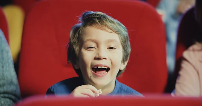 Portrait of the cute little Caucasian blond boy watching comedy movie in the cinema and laughing joyfully. Close up.