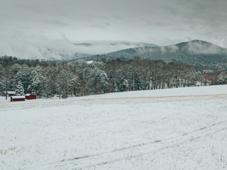 A landscape, horizontal frame of a country field and red home after a severe winter storm. Storm clouds break up over the mountains displaying beautiful snow covered fields, trees, and mountains.