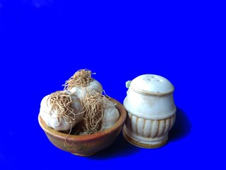 Garlic heads over clay bowl with salt shaker on blue background