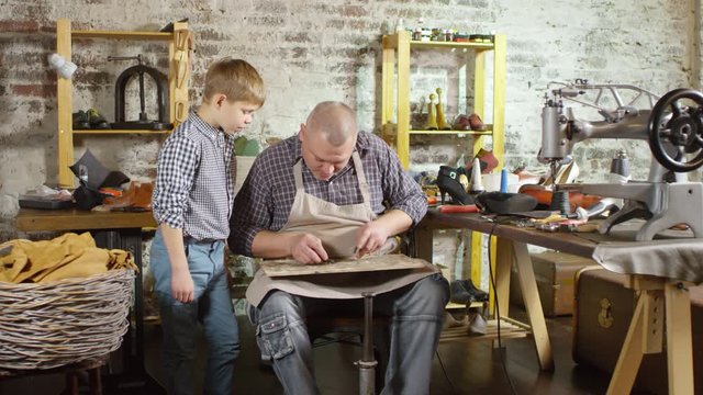 Professional shoemaker explaining and showing his little son how to cut heel tip and repair female boot while working in the workshop