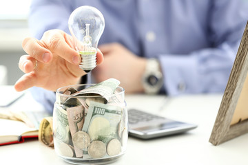 Male arm holding lightbulb over tip can full of US money closeup
