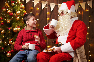 Santa Claus and child boy drinking tea, eating cookies, talking and sitting indoor near decorated xmas tree with lights - Merry Christmas and Happy Holidays!