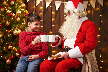 Santa Claus and child boy drinking tea, eating cookies, talking and sitting indoor near decorated xmas tree with lights - Merry Christmas and Happy Holidays!