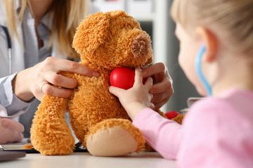 Little girl hold in arms toy red hear playing with teddy bear while visiting doctor