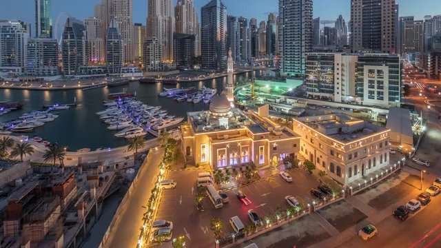 Yachts in Dubai Marina flanked by the Al Rahim Mosque and residential towers and skyscrapers aerial night to day transition timelapse. Modern high rise skyline with boats and wooden dhows