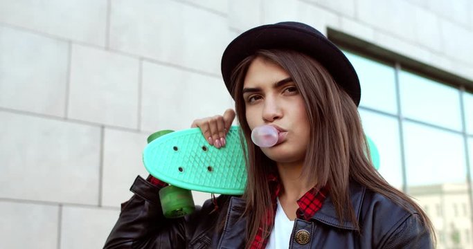 Portrait of the beautiful young Caucasian girl in hipster style and black hat holding skateboard on her shoulder, looking at the camera and blowing bubble gum with serious face Outdoors. Close up.