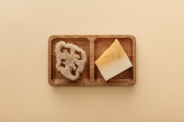 top view of brown wooden soapdish with loofah and piece of soap on beige background