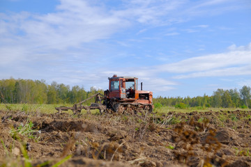 A tracked tractor with a plough plows the ground in autumn against the background of the forest.