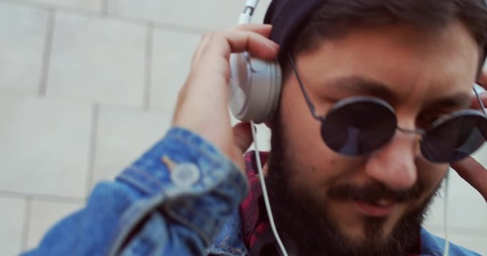 Close up of the young hipster stylish Caucasian man in dark glasses putting on headphones and listening to the music. Outdoor.