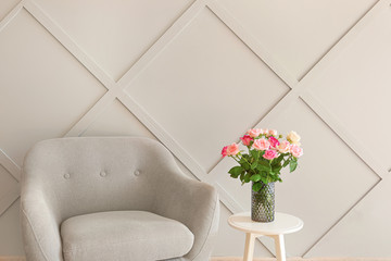 Beautiful rose flowers in vase on table and armchair near grey wall in room