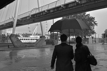 Black and white of unidentifiable couple with umbrella in the rain in London with Millenium Bridge and London Eye in background