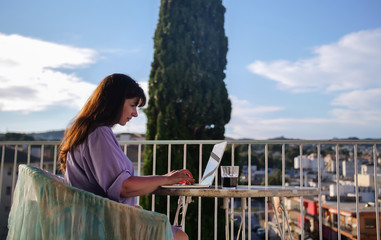 Brunette girl uses her laptop while sitting on the balcony in summer