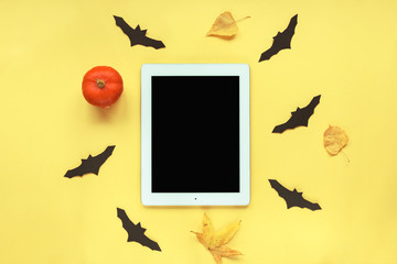 Tablet with blank screen and pumpkin, autumn leaves, handmade black bats on yellow background. Halloween concept. Top view, flat lay.